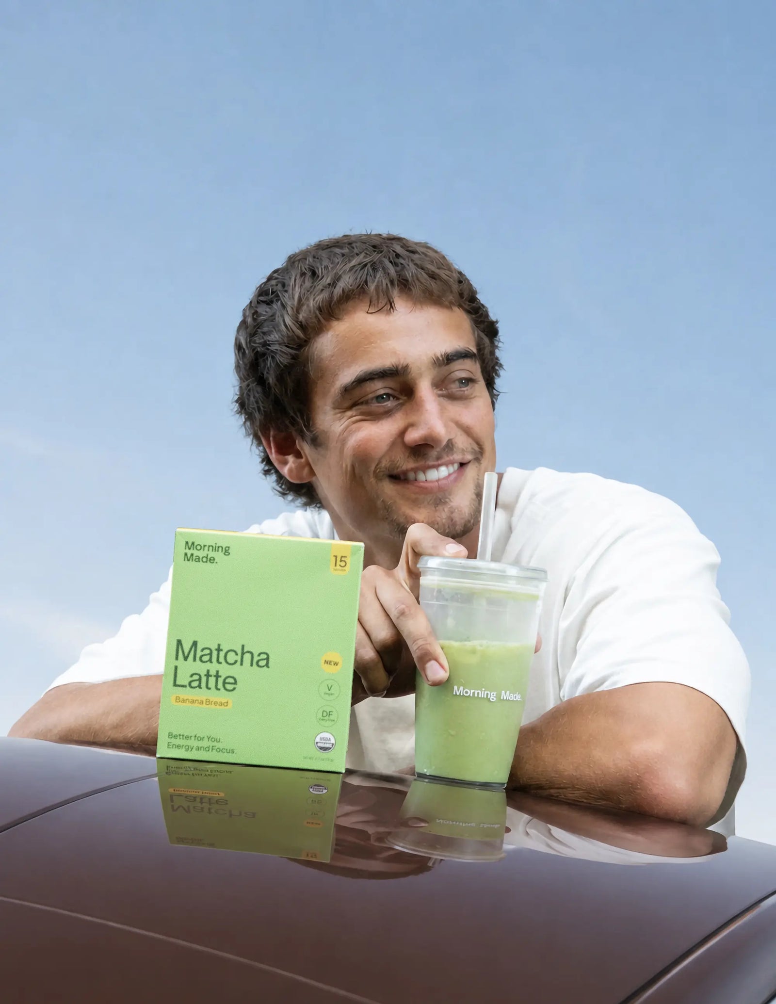 A young man in a white T-shirt smiles off-camera while leaning on the glossy surface of a car against a blue sky. In front of him sits a light green box of "Morning Made. Matcha Latte" in Banana Bread flavor, alongside a clear plastic cup filled with iced green matcha latte and a straw.
