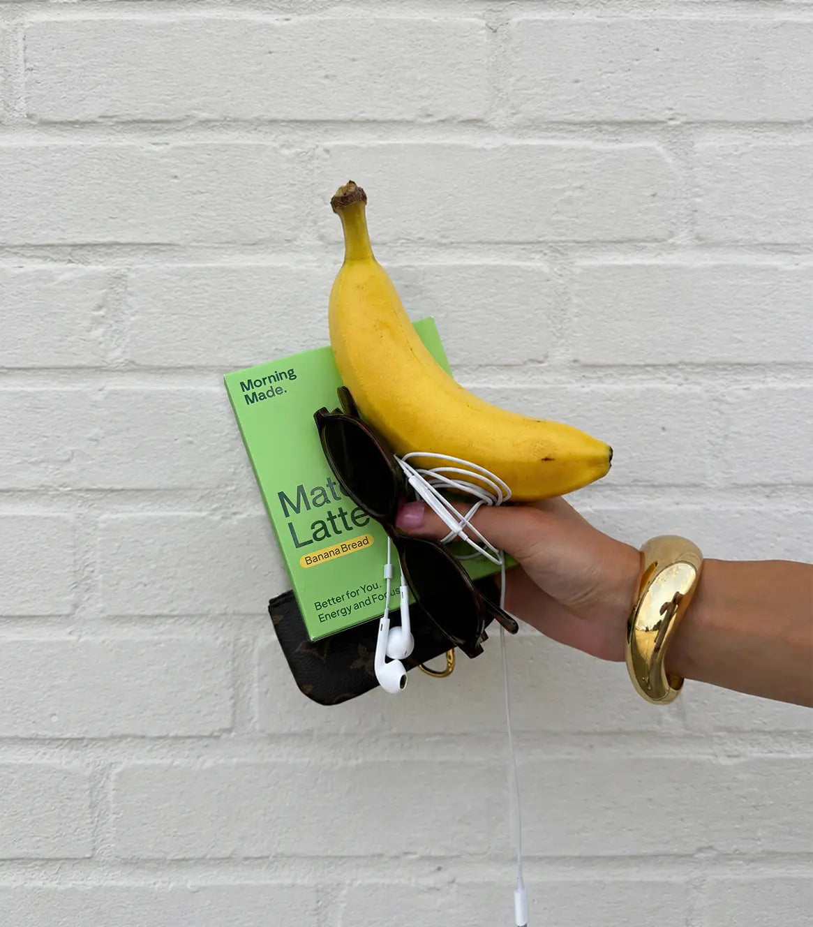 A hand with a thick gold cuff bracelet holds an assortment of items against a white painted brick wall. The items include a green box of "Morning Made. Matcha Latte" Banana Bread flavor, a fresh yellow banana resting on top, dark sunglasses, white wired AirPods, and a small dark designer key pouch underneath.
