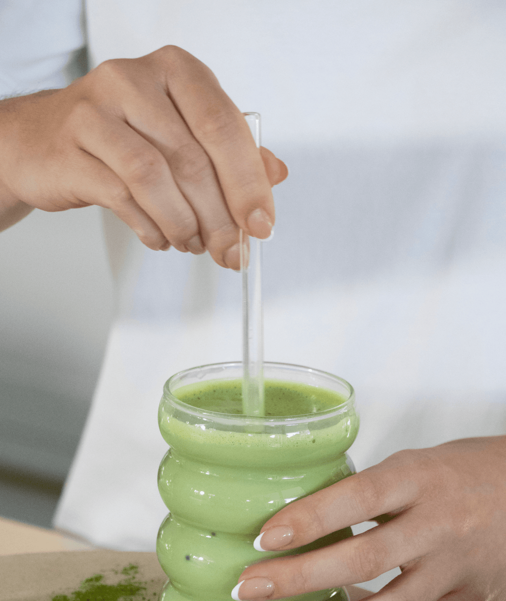 Person wearing a white t-shirt holding a glass full of matcha with a visible clear glass straw sitting in the glass