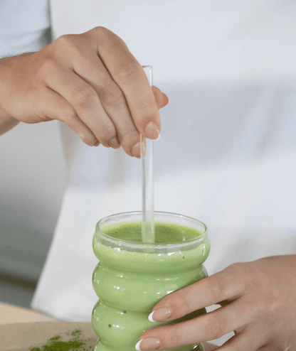 Person wearing a white t-shirt holding a glass full of matcha with a visible clear glass straw sitting in the glass